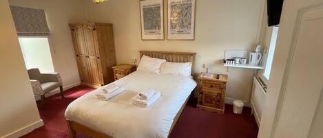 A neatly arranged hotel room with a large bed, a wooden wardrobe, a small bedside table with a lamp, and a window with a view.