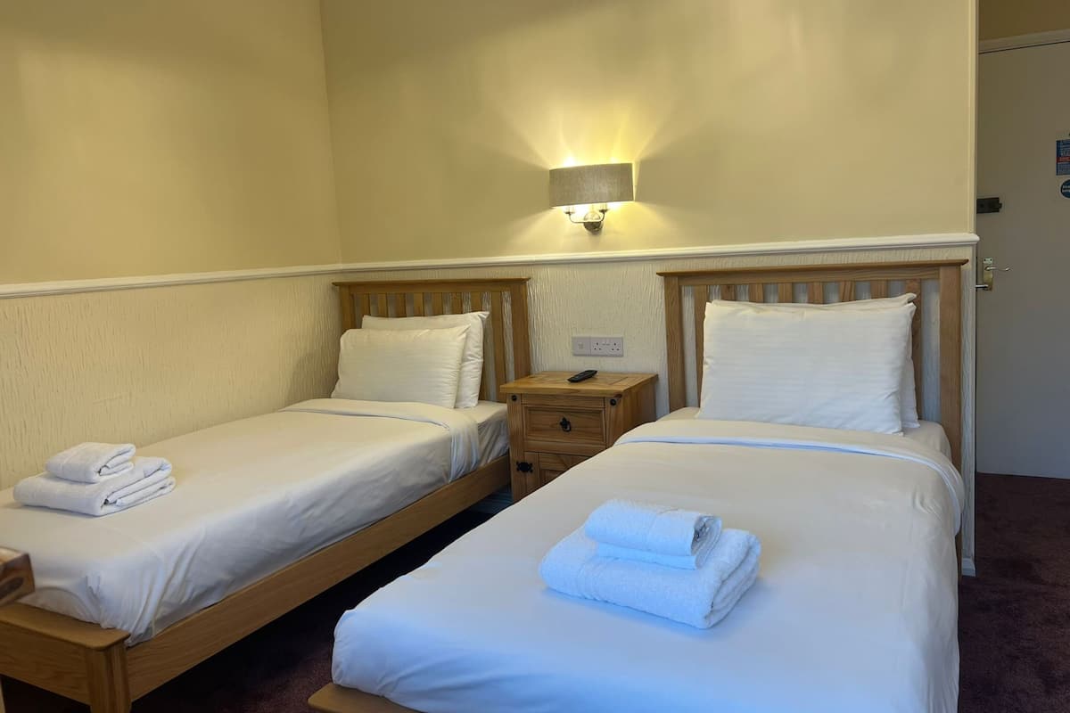 A hotel room with two single beds, each with white linens and pillows, a wooden nightstand with a remote control, and a wall-mounted light fixture.