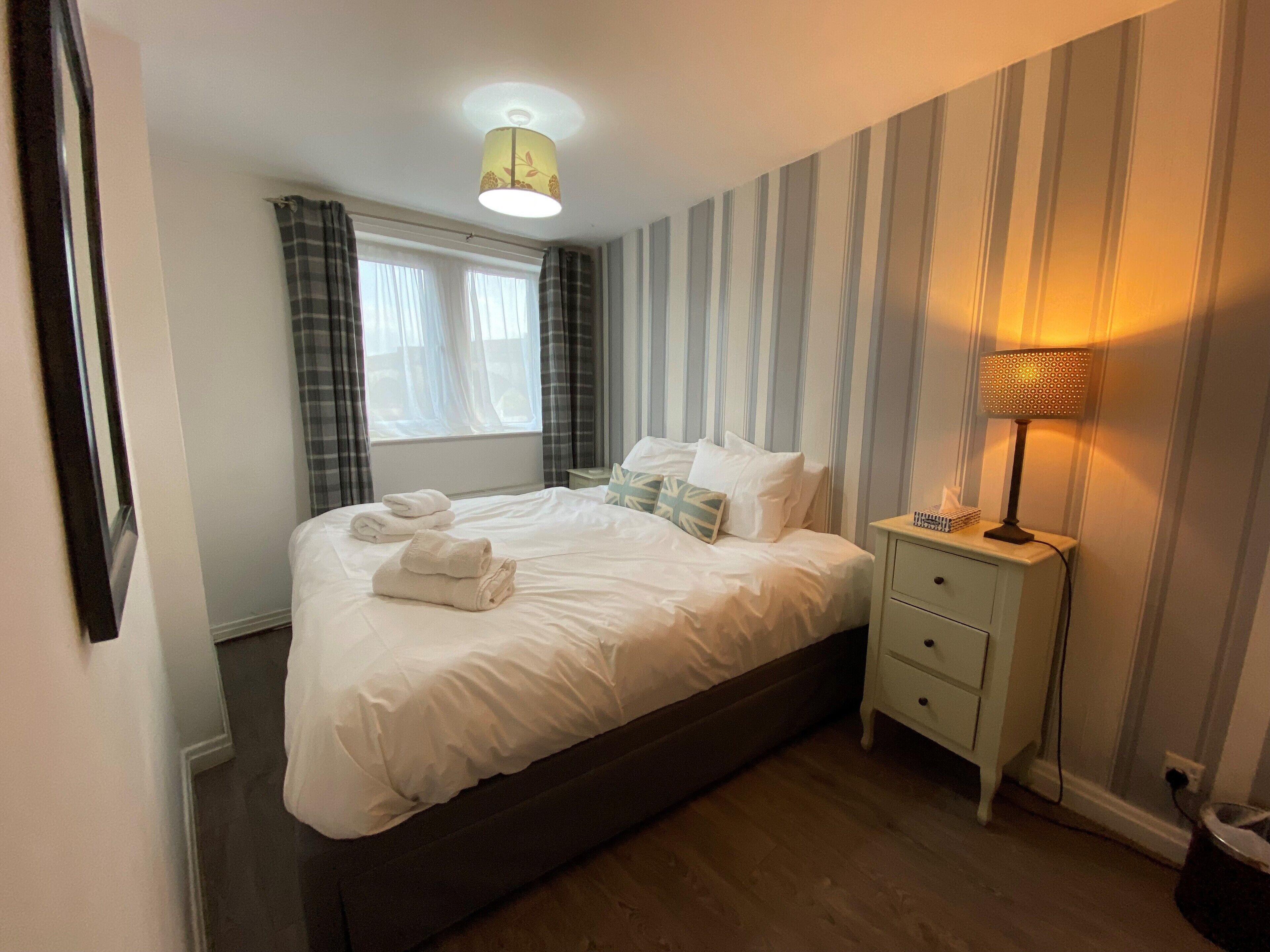 A neatly made bed with white linens and a pillow with a leaf pattern, a bedside lamp, and a window with curtains.