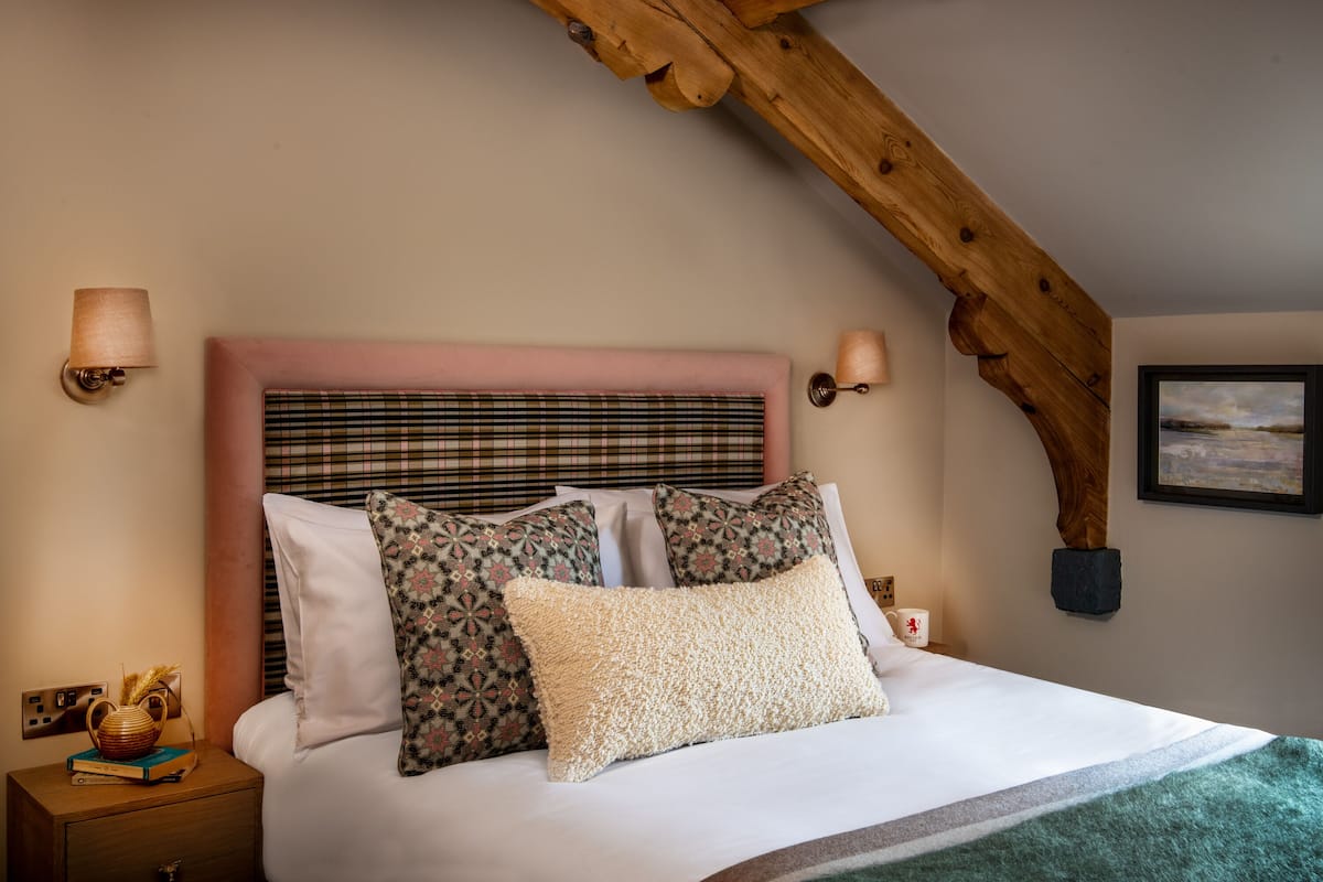 A neatly made bed with patterned pillows, a bedside table with a book and a vase, and a wooden ceiling beam.