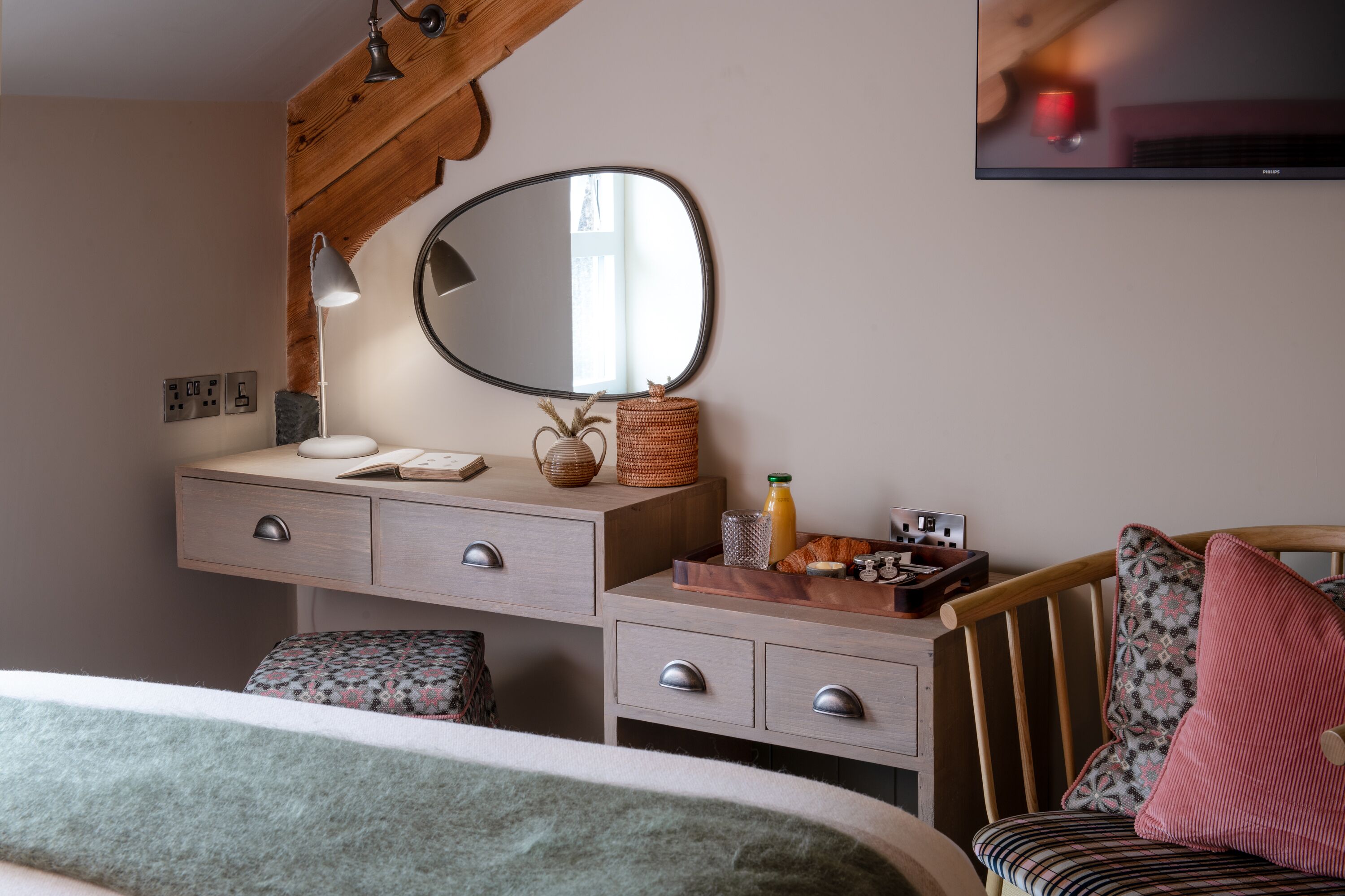 A bedroom with a wooden headboard, a bedside table with a lamp, a mirror, and a TV mounted on the wall.