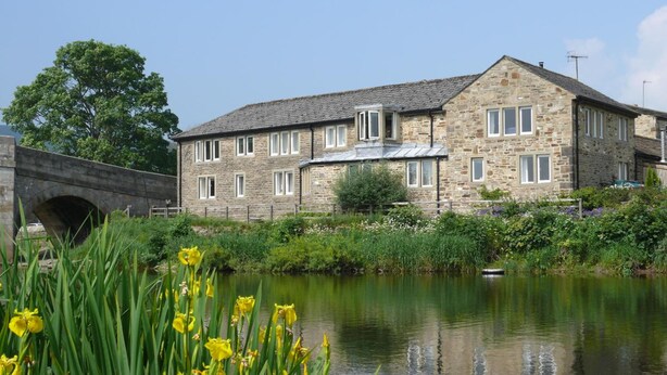 A stone building with multiple windows and a sloped roof, situated next to a calm river with yellow flowers in the foreground. - Red Lion Hotel and Manor House (Skipton)