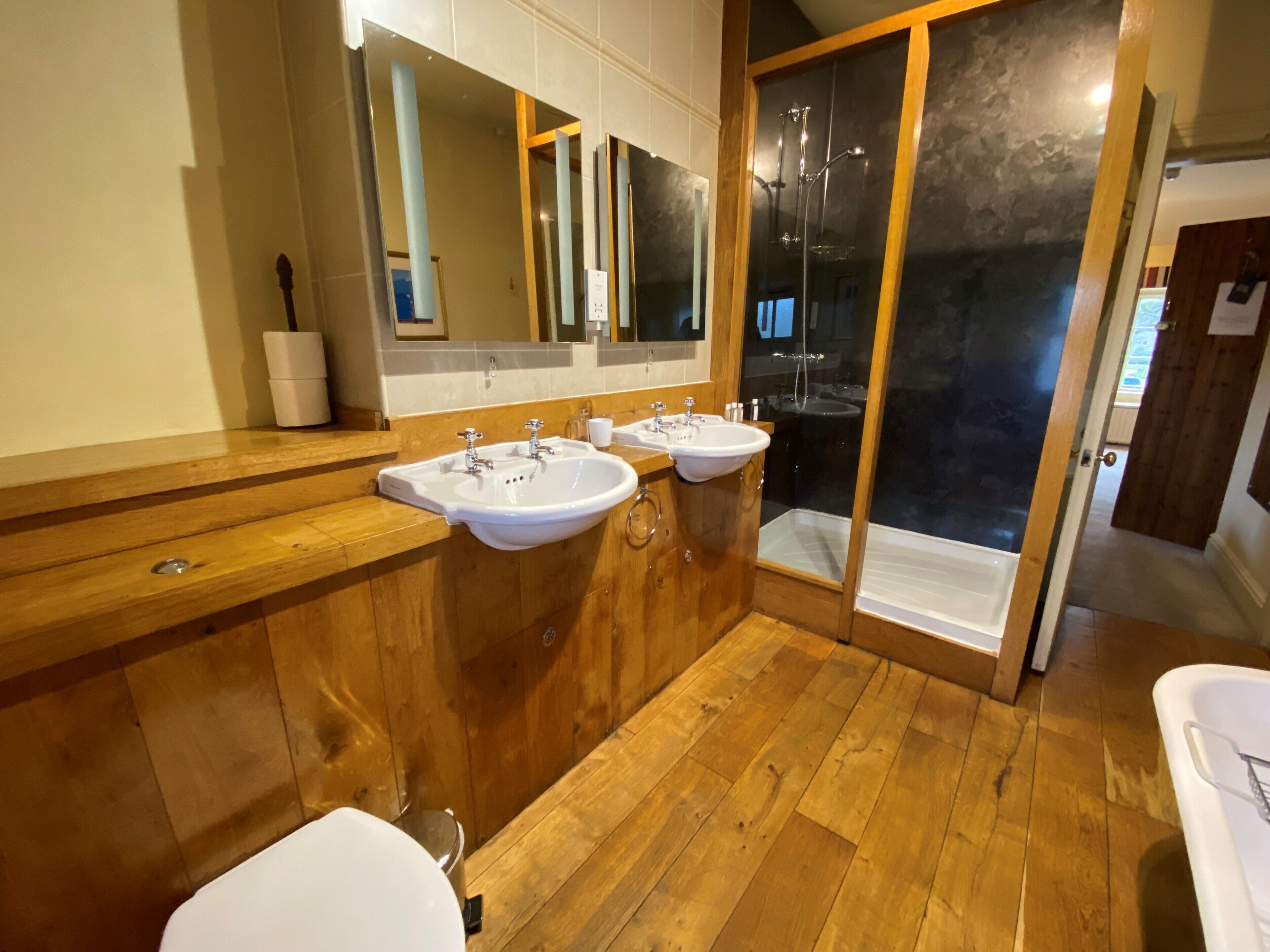 Ein modernes Badezimmer mit Holzboden, zwei Waschbecken, einem großen Spiegel und einer Duschkabine.