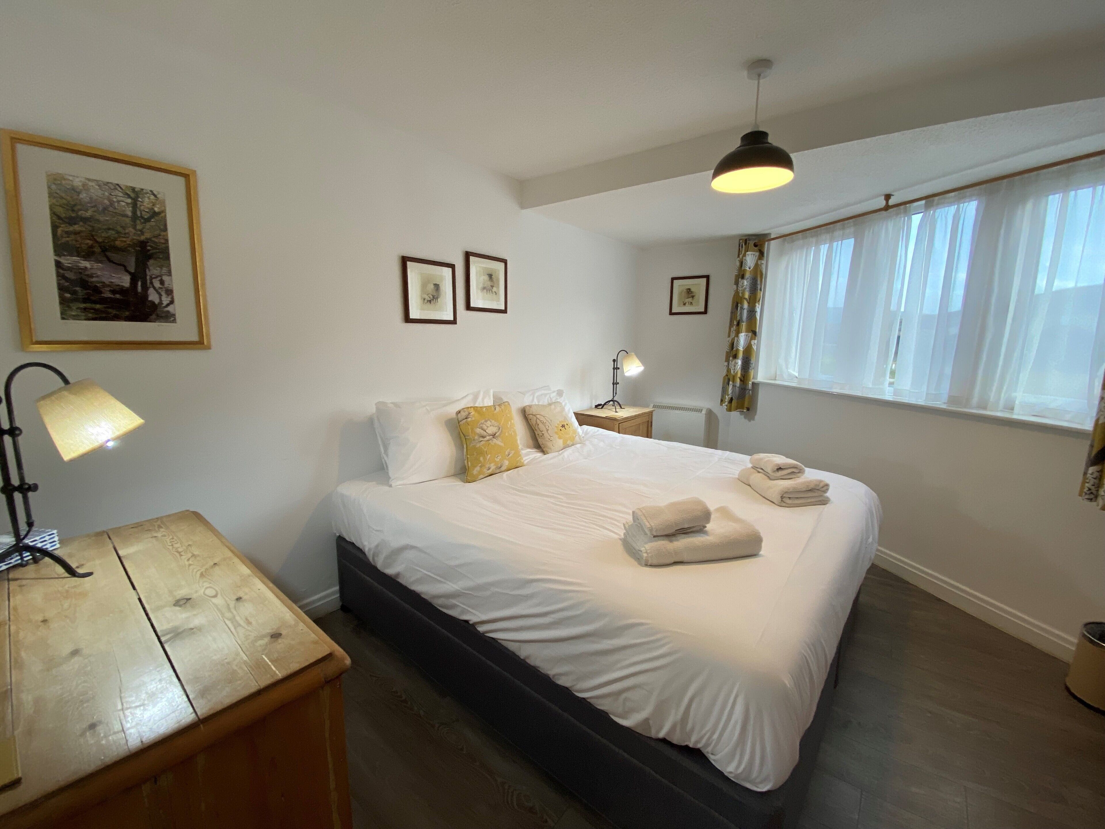 Un lit bien fait, avec du linge de lit blanc et quelques coussins décoratifs, une table de chevet avec une lampe et une fenêtre ornée de rideaux légers.