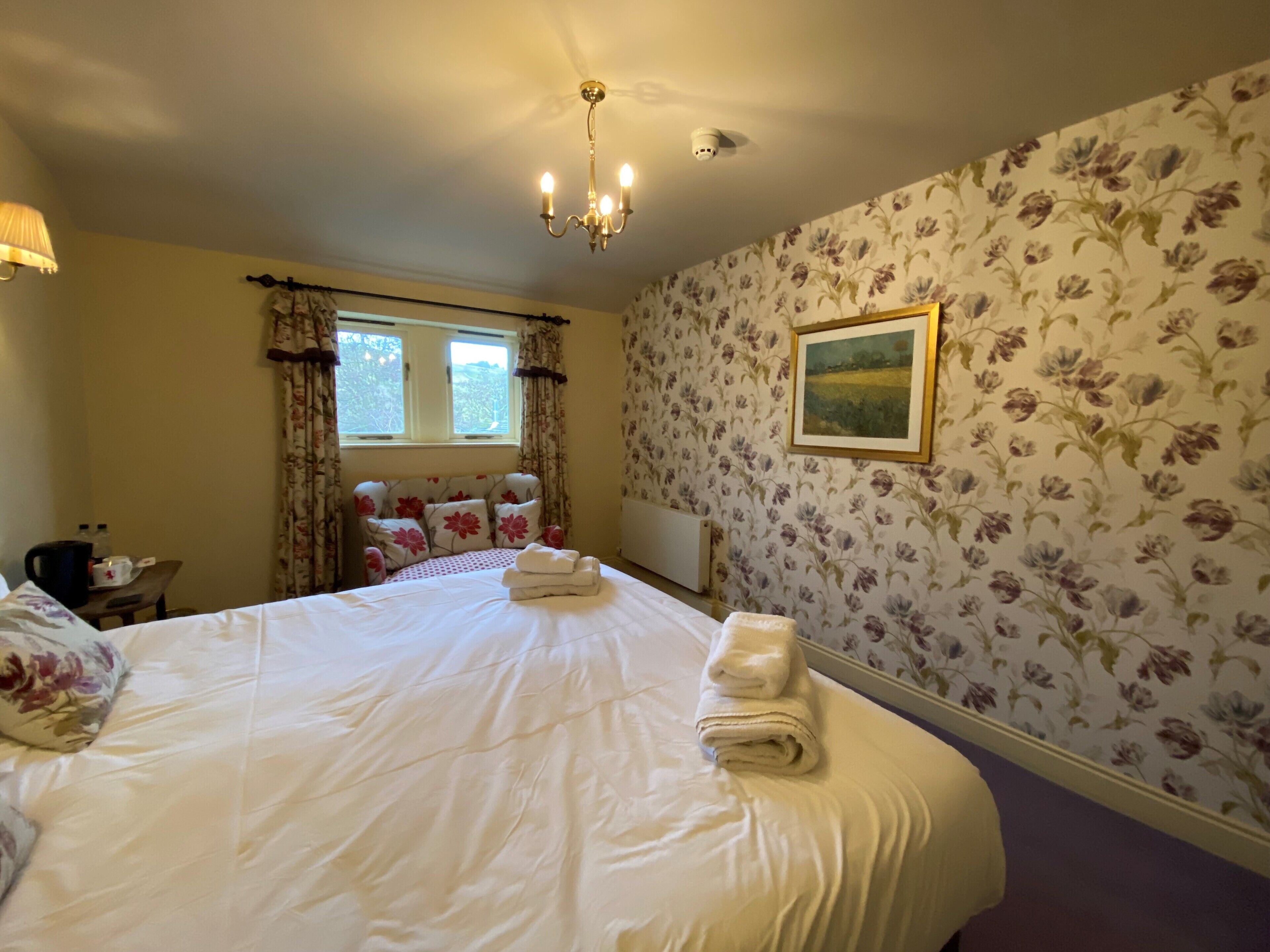 A hotel room with a large bed, floral wallpaper, a chandelier, and a painting on the wall.