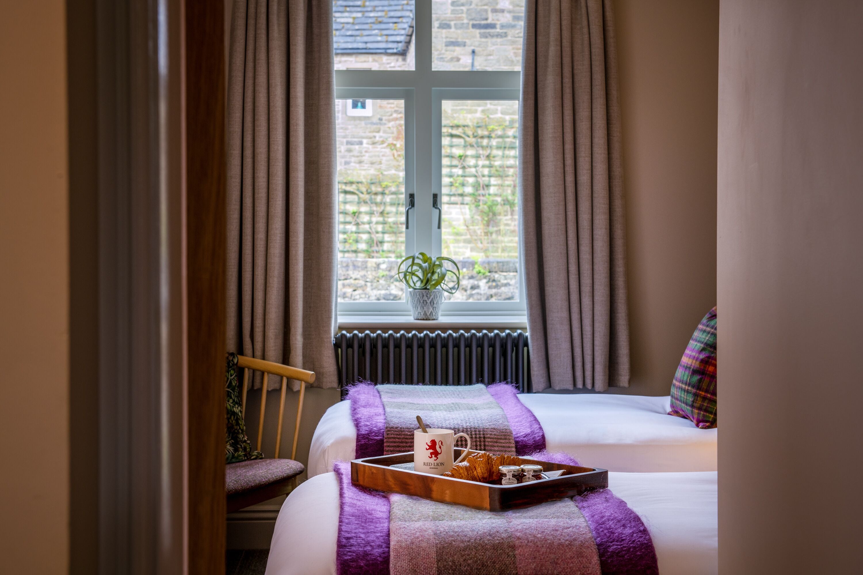 Ein Schlafzimmer mit Bett, einem Fenster mit Vorhängen, einem Stuhl und einem Tablett mit Gegenständen auf dem Bett.