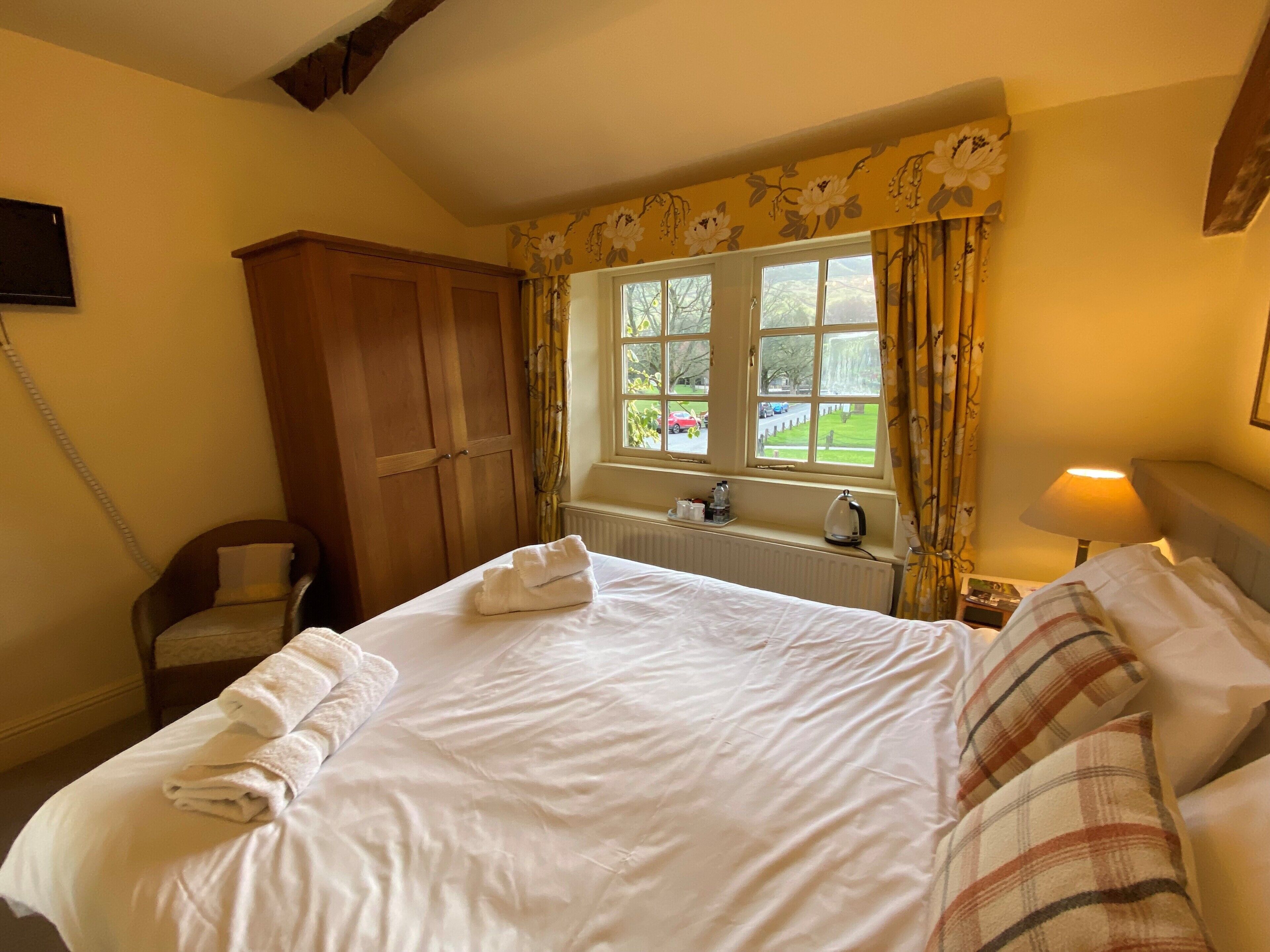 Una camera d'albergo con un letto grande, un armadio in legno, una finestra con tende, un termosifone e un comodino con una lampada.