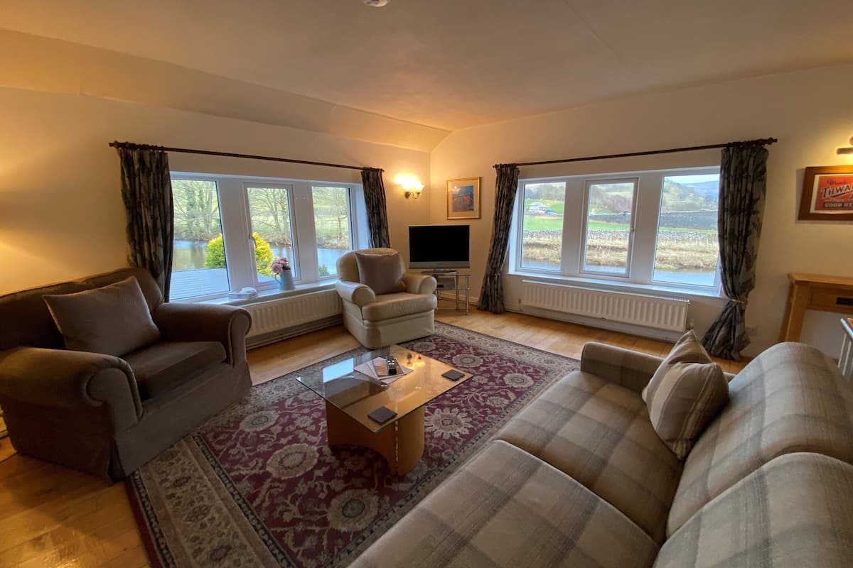 A spacious living room with a large sofa, a smaller armchair, and a coffee table. There are two windows with curtains, a flat-screen TV, and a fireplace.