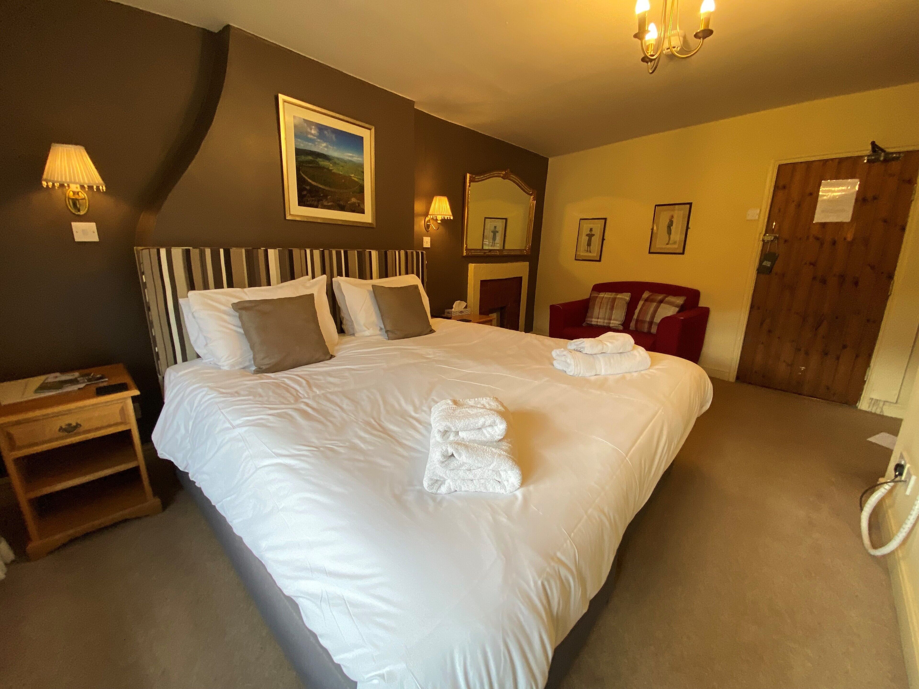 A hotel room with a large bed, a wooden nightstand, a red armchair, a wooden door, and a painting on the wall.
