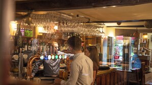 A bar area with a bartender preparing drinks, a woman standing by the counter, and a variety of bottles and glasses on shelves. - Red Lion Hotel and Manor House (Skipton)