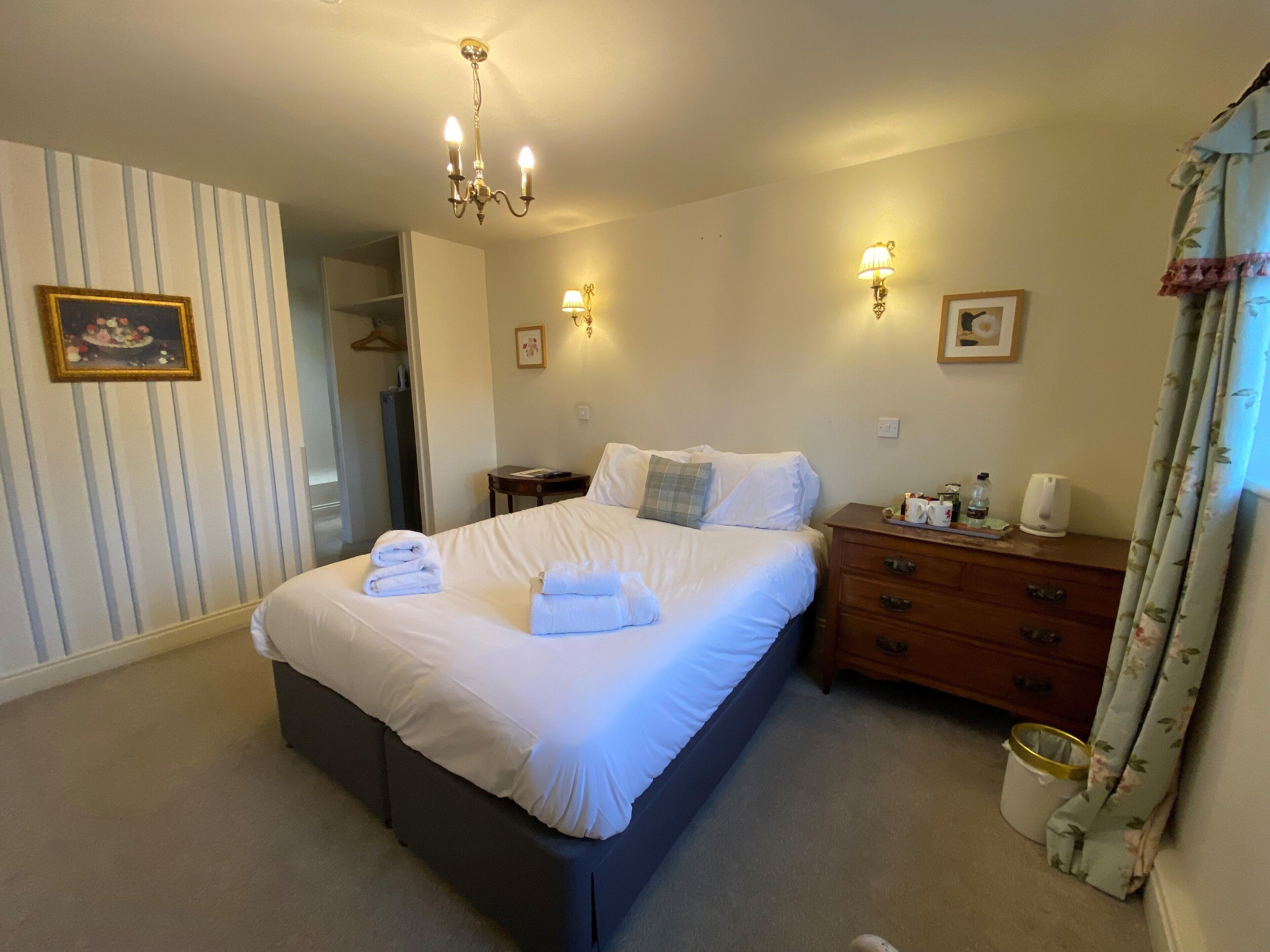 A neatly made bed with white linens and towels, a wooden dresser with drawers, a small table with a lamp, and a chandelier.