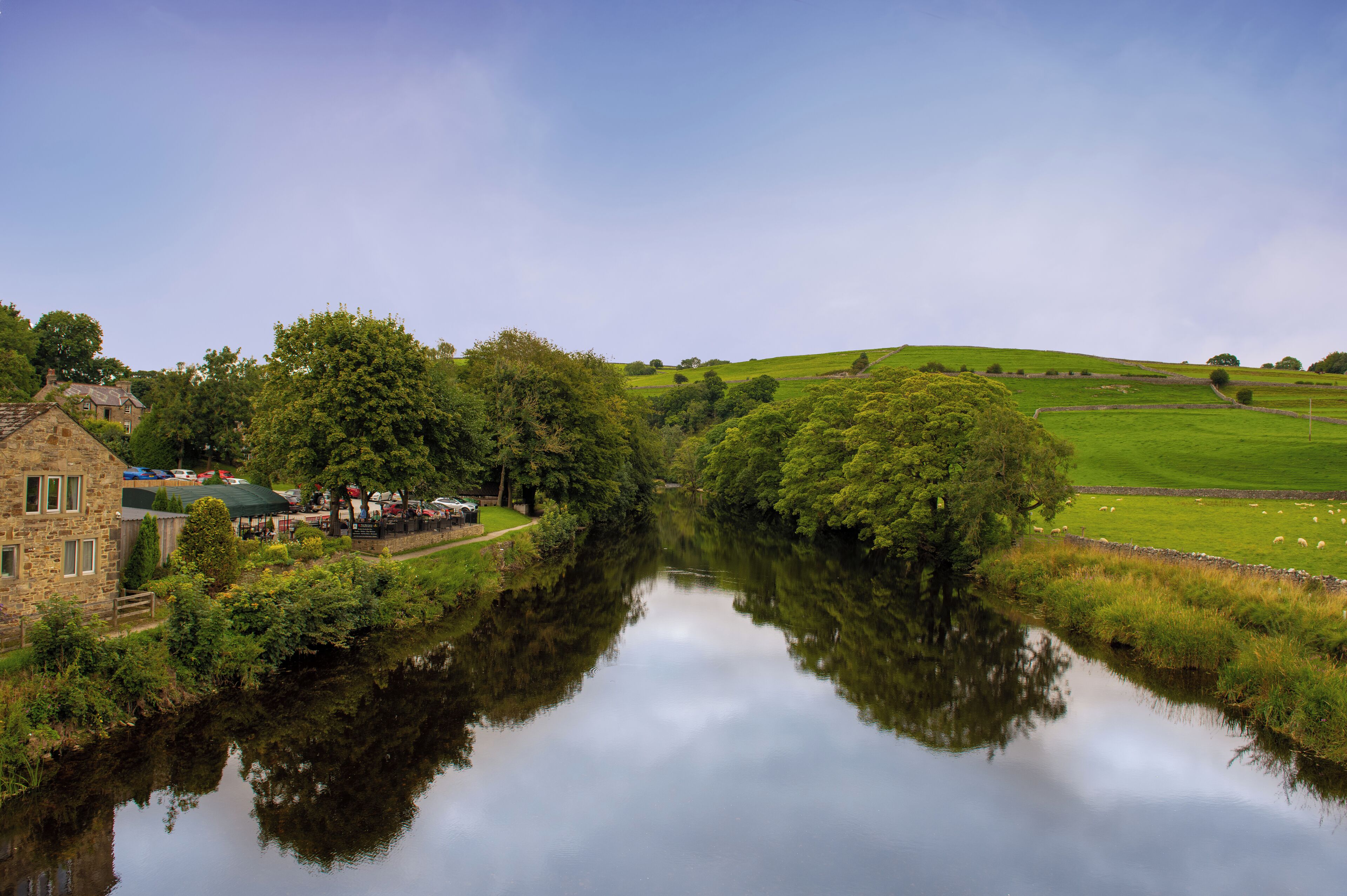 A river reflecting the surrounding landscape, a stone building, and a grassy field with sheep.