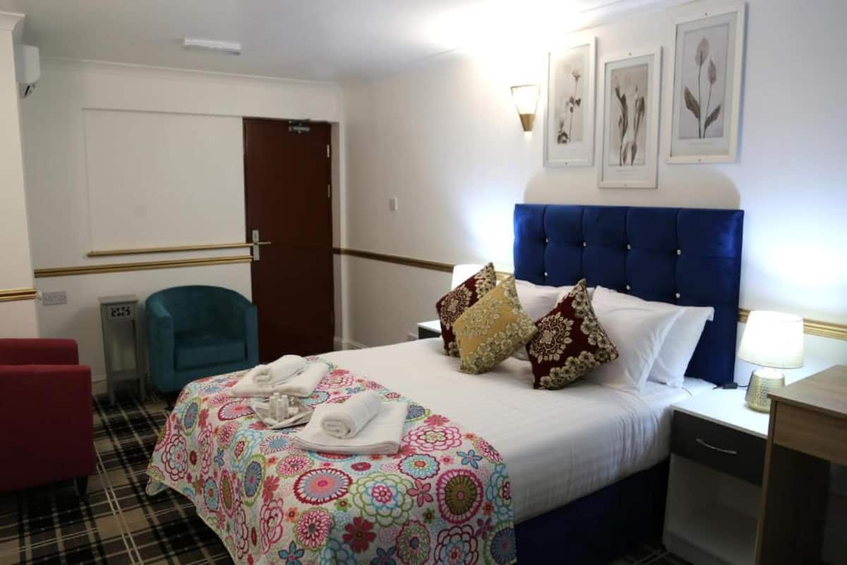 A hotel room with a bed, a blue headboard, a bedside table with a lamp, a chair, and a door.
