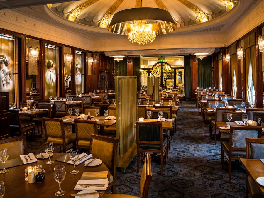 Un restaurante espacioso con numerosas mesas y sillas, candelabros elegantes y una alfombra con diseño.