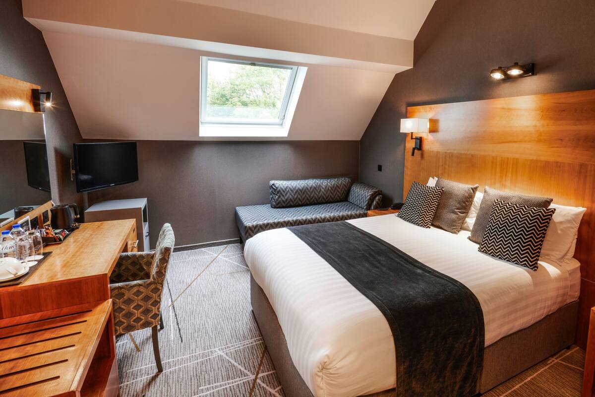 A modern hotel room with a large bed, a desk with a chair, a flat-screen TV, and a skylight.