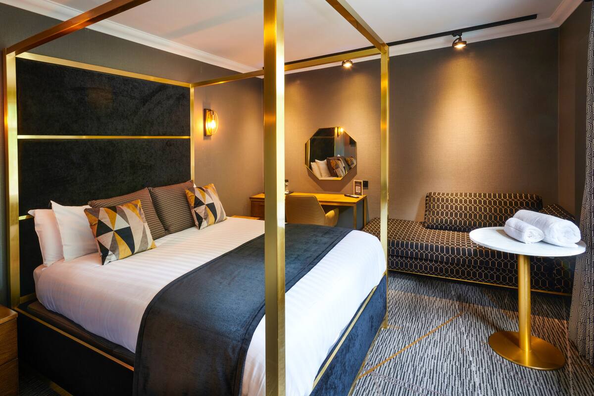A four-poster bed with a dark headboard, a bedside table, a small desk with a chair, a round table with a towel, and a patterned sofa.