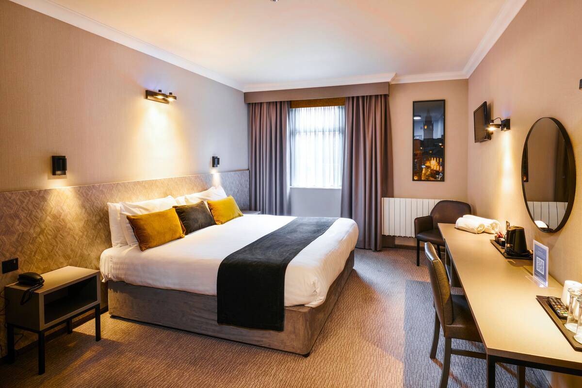 A hotel room with a large bed, bedside tables, a desk with a chair, a mirror, and a window with curtains.