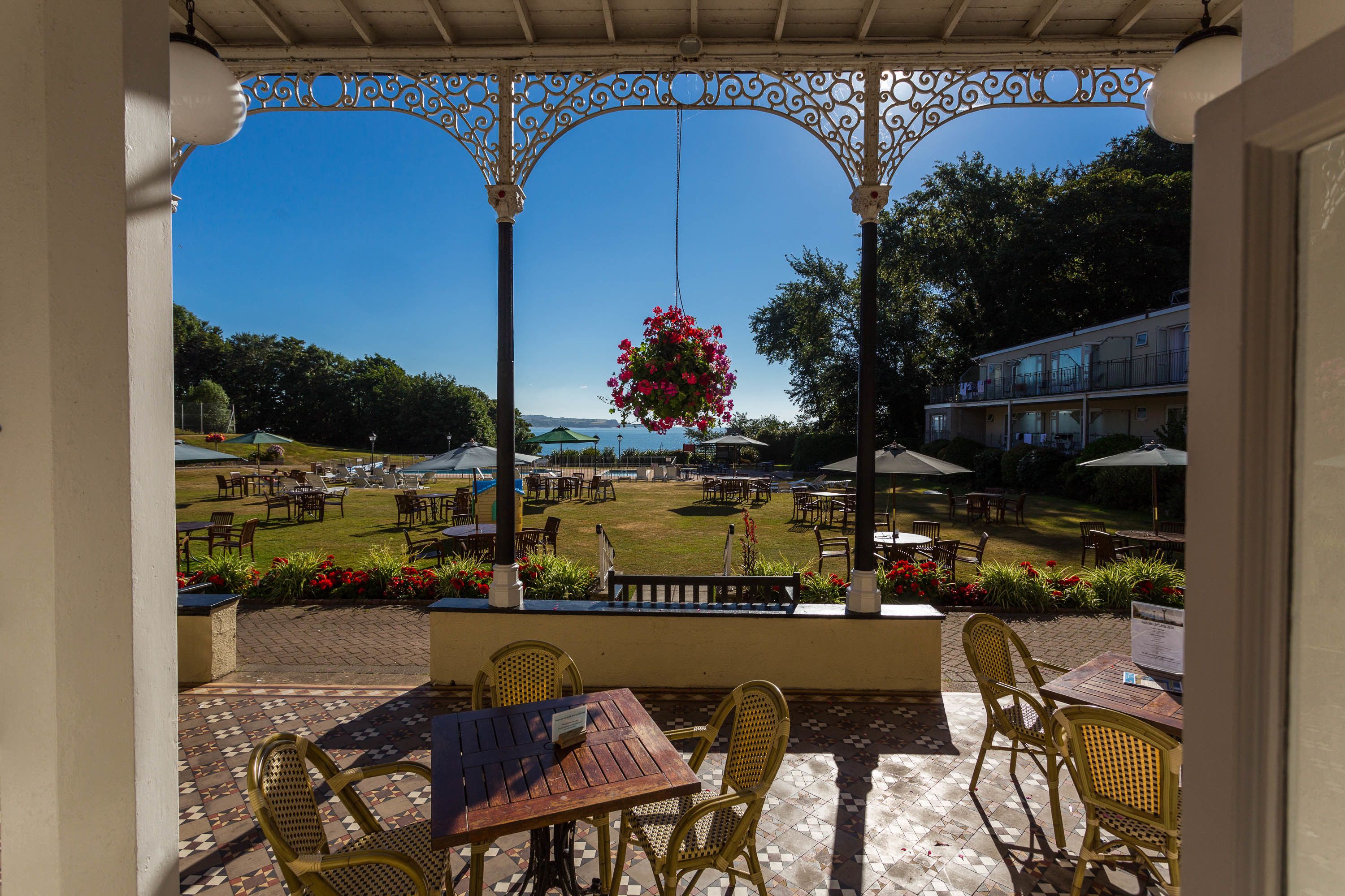 An outdoor patio with wicker chairs and tables, a hanging flower basket, and a view of a lawn and buildings in the background.