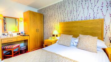A hotel room with a large bed, wooden wardrobe, bedside table with lamp, and a small stool.