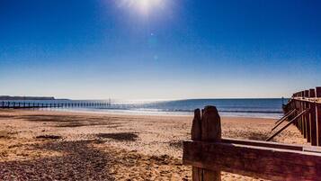 A beach with a wooden pier extending into the water, a clear sky, and the sun shining brightly.