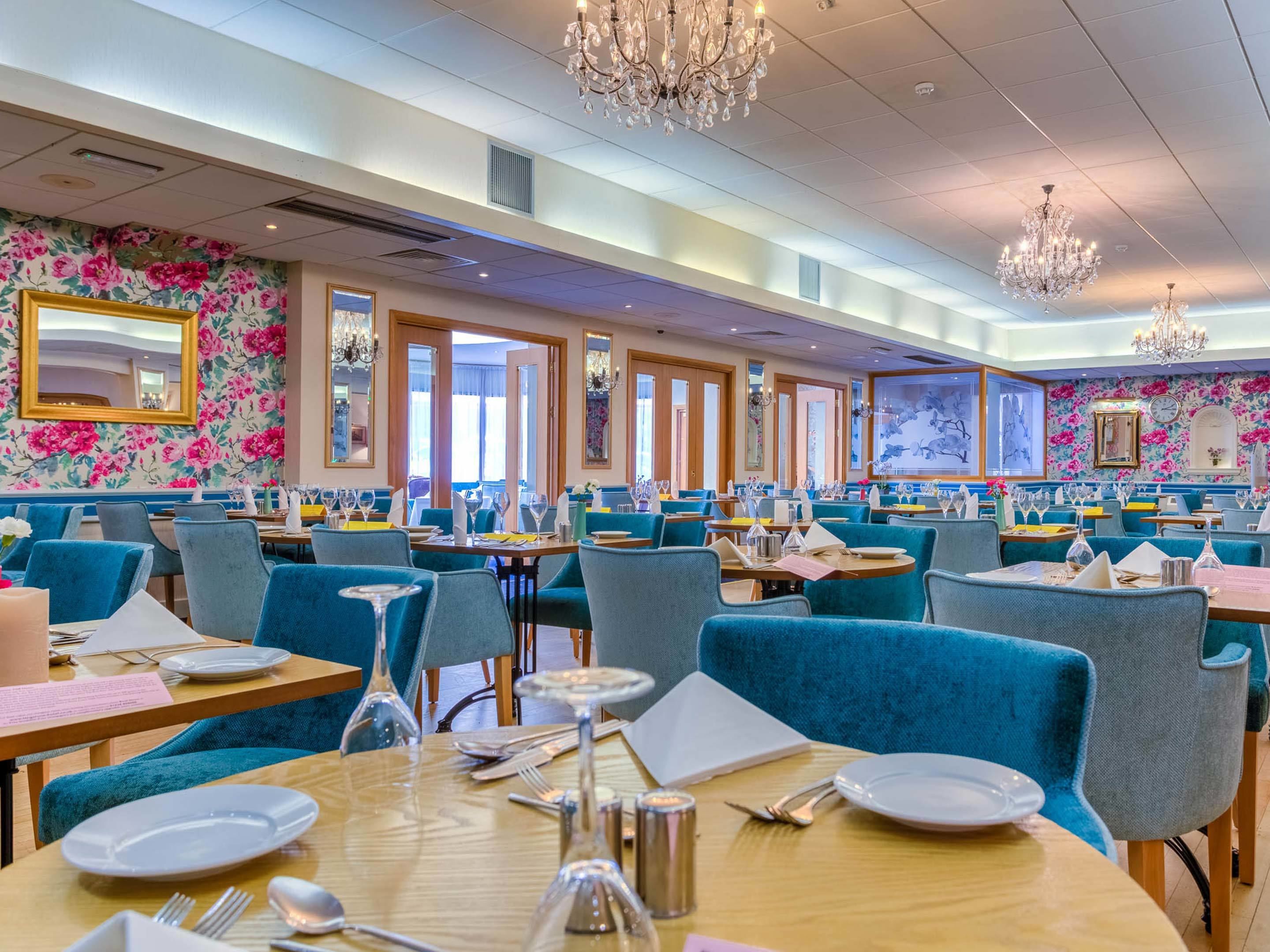 A spacious restaurant with blue upholstered chairs, wooden tables, and floral wallpaper.