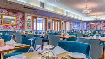 A spacious restaurant with blue upholstered chairs, wooden tables, and floral wallpaper.