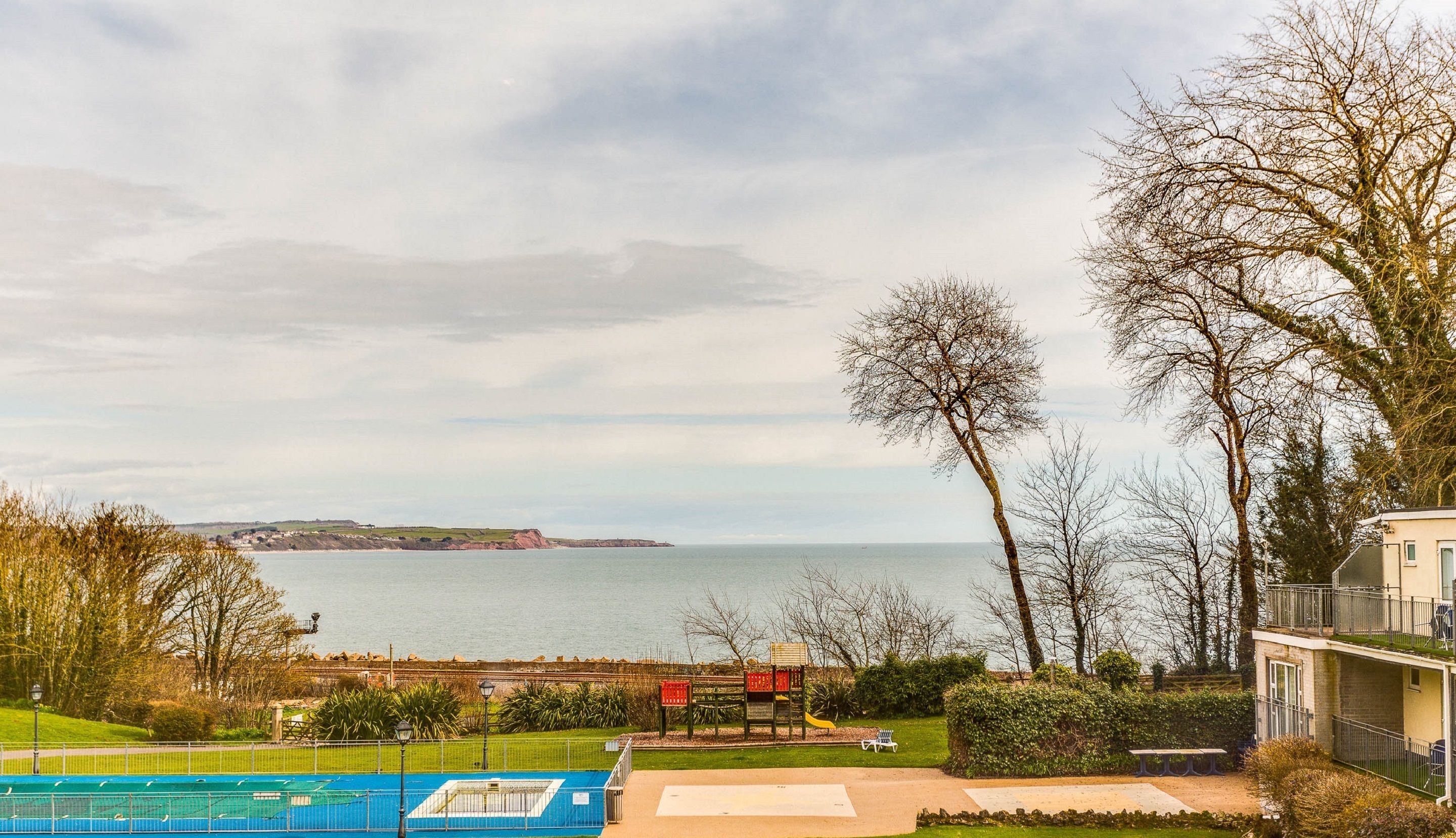 A coastal view with a swimming pool, a playground, and a building with a balcony overlooking the sea.