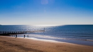 Una playa con un muelle de madera que se adentra en el agua, un cielo despejado y un mar en calma.