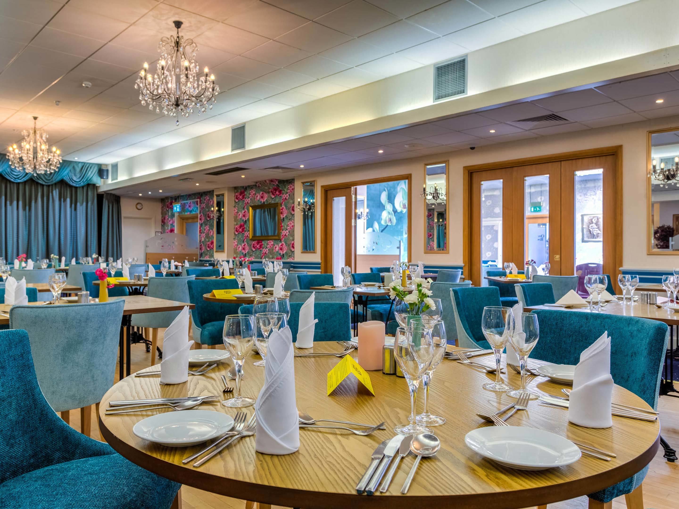 Un restaurant avec des tables rondes dressées pour le service, doté de chaises bleues rembourrées et de lustres.