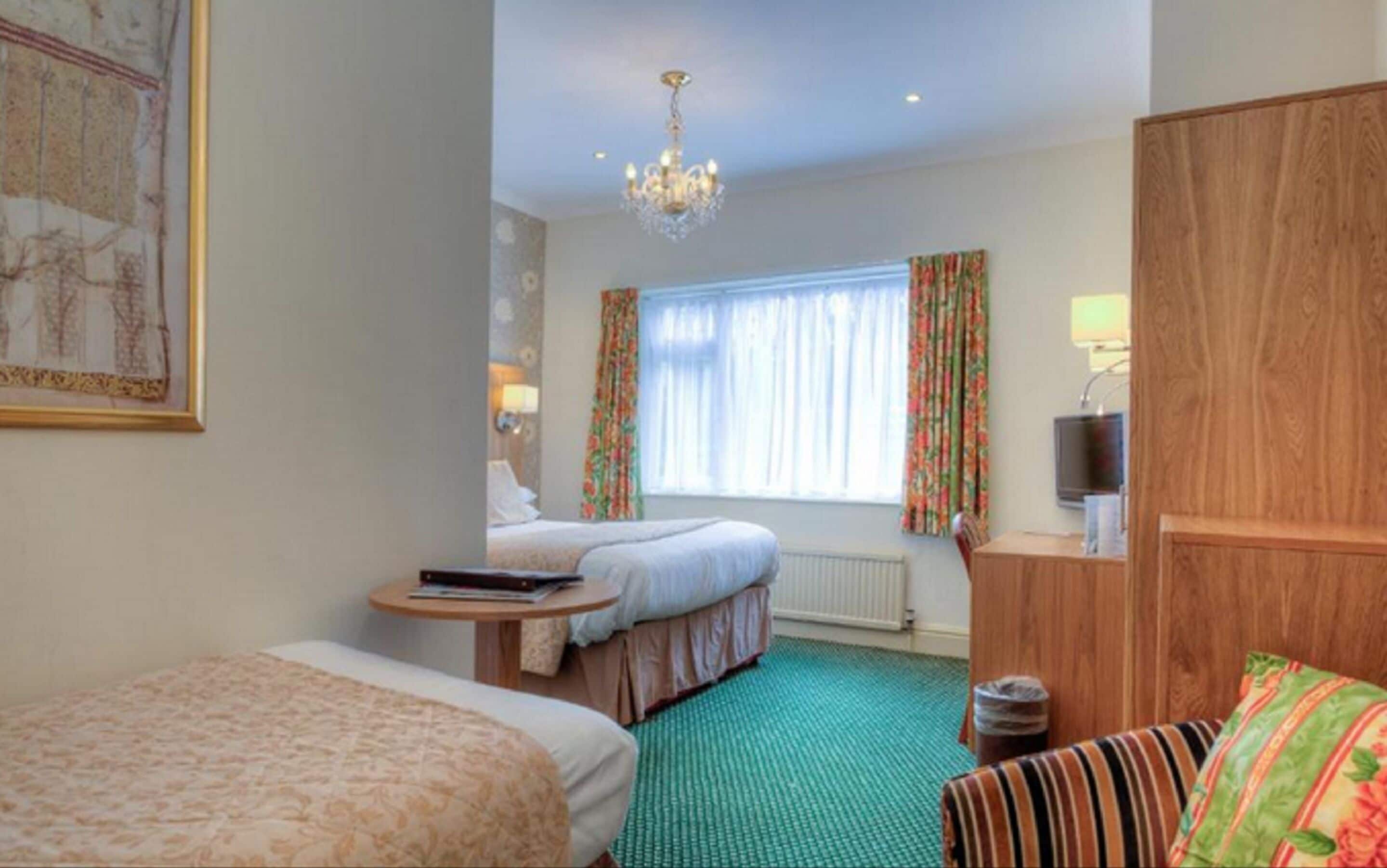 A hotel room with two beds, a green carpet, a chandelier, a television, and a wooden wardrobe.
