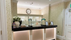 A reception desk with a clock above it, a vase with flowers, and a framed certificate on the counter. - Athena (London)