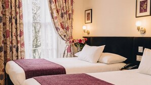 A hotel room with two beds, a nightstand with a phone, wall-mounted lights, and a window with curtains. - Athena (London)