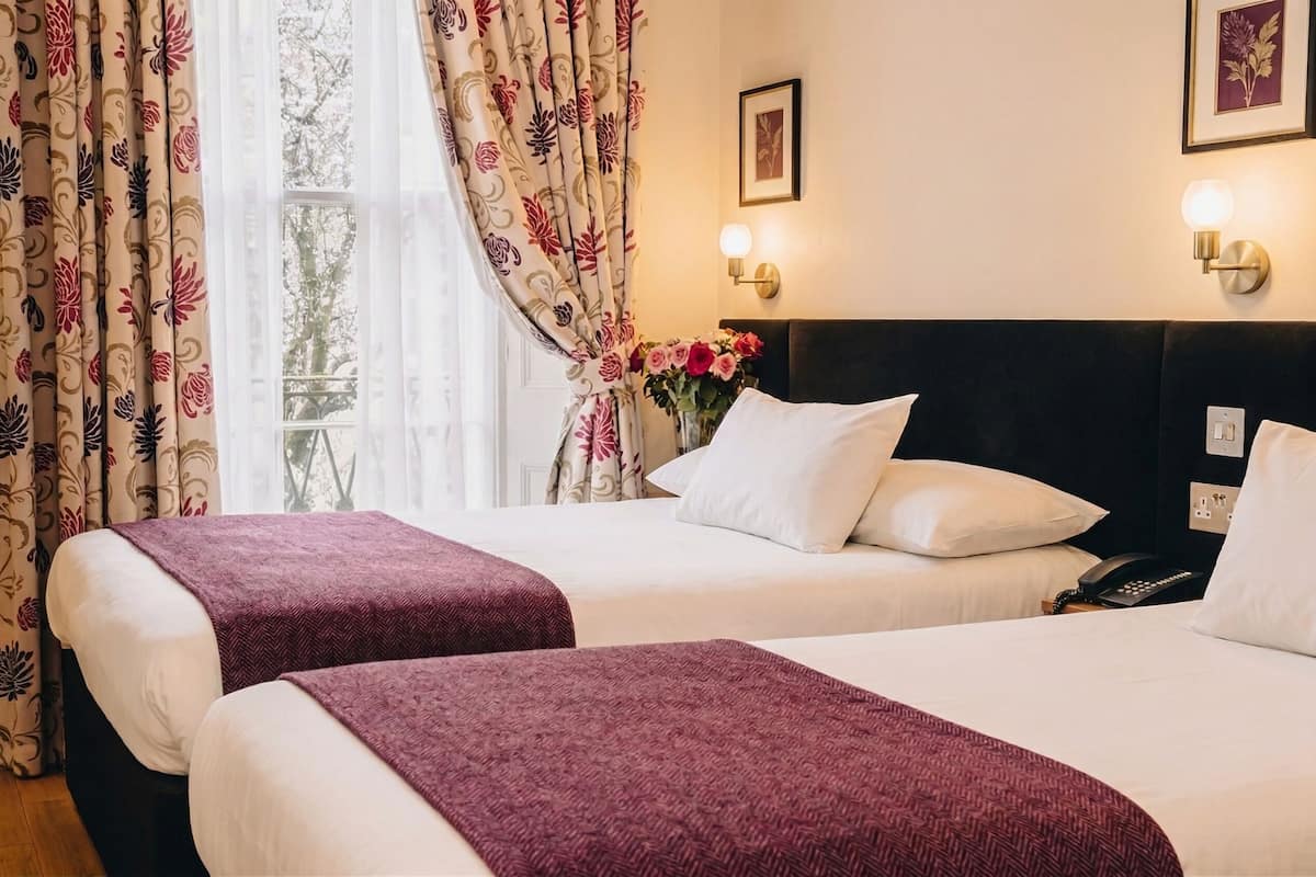 A hotel room with two beds, a nightstand with a phone, wall-mounted lights, and a window with curtains.