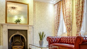A room with a fireplace, a red leather sofa, a glass coffee table, and floral curtains. - Athena (London)