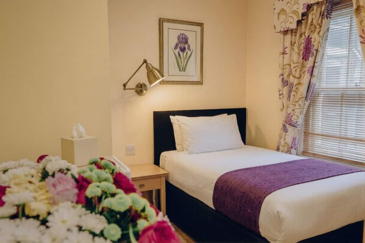 A hotel room with a bed, bedside table, lamp, framed artwork, and a window with curtains.