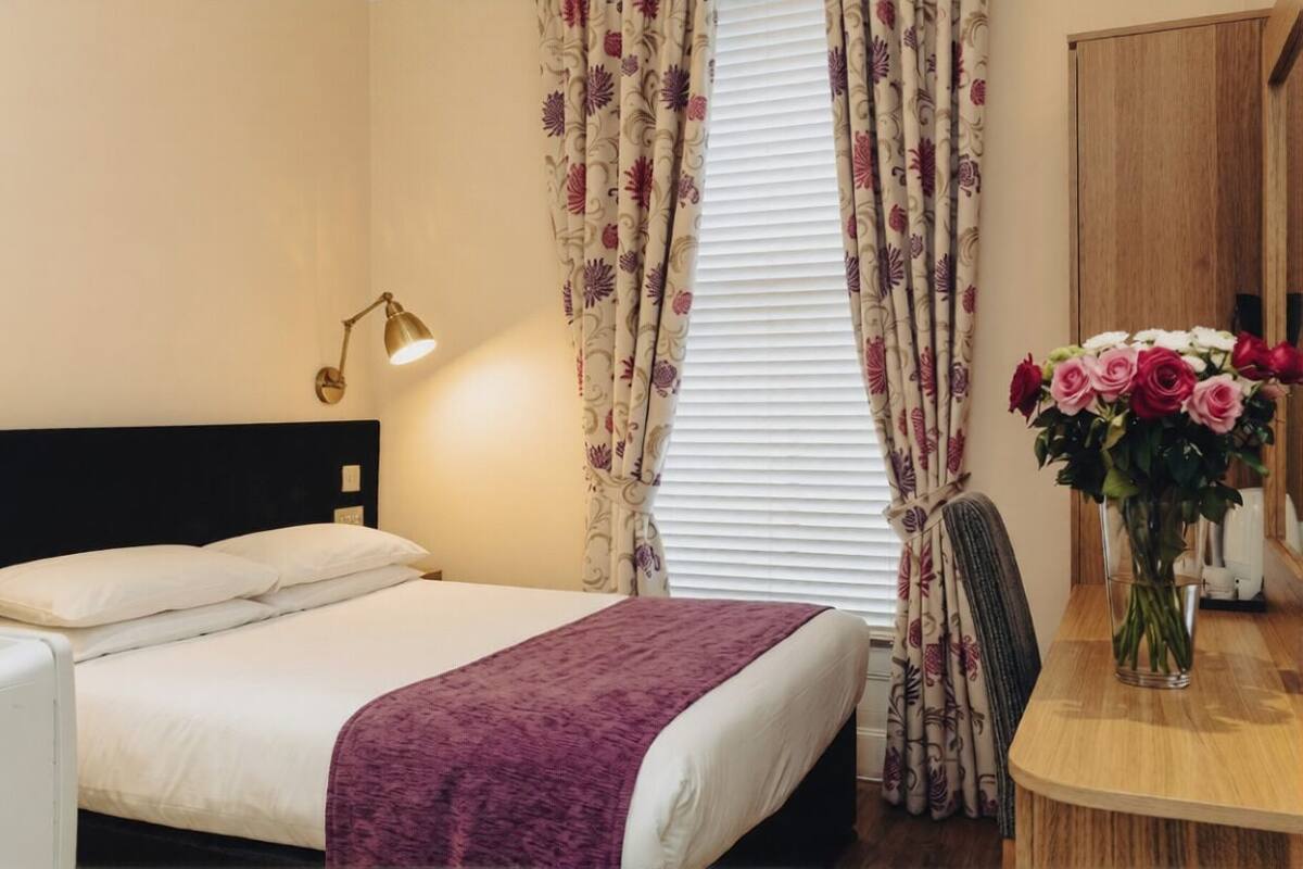 A hotel room with a bed, a bedside lamp, a window with curtains, and a vase with flowers on a table.