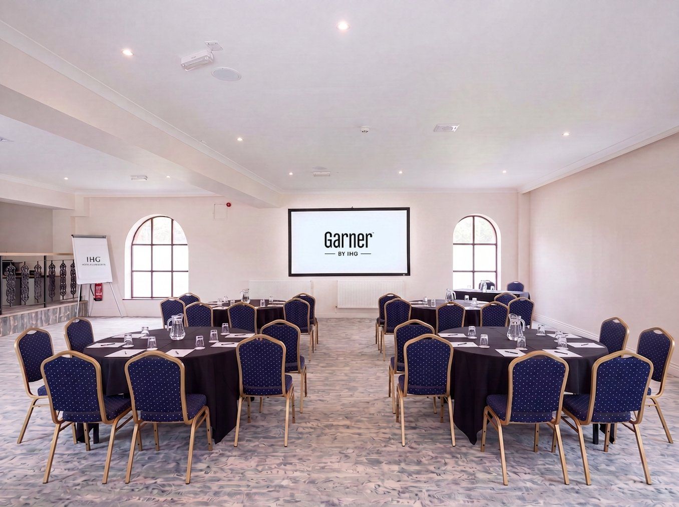 A conference room with multiple tables and chairs, a projector screen displaying 'Garnier by IHG', and a whiteboard with 'IHG' on it.