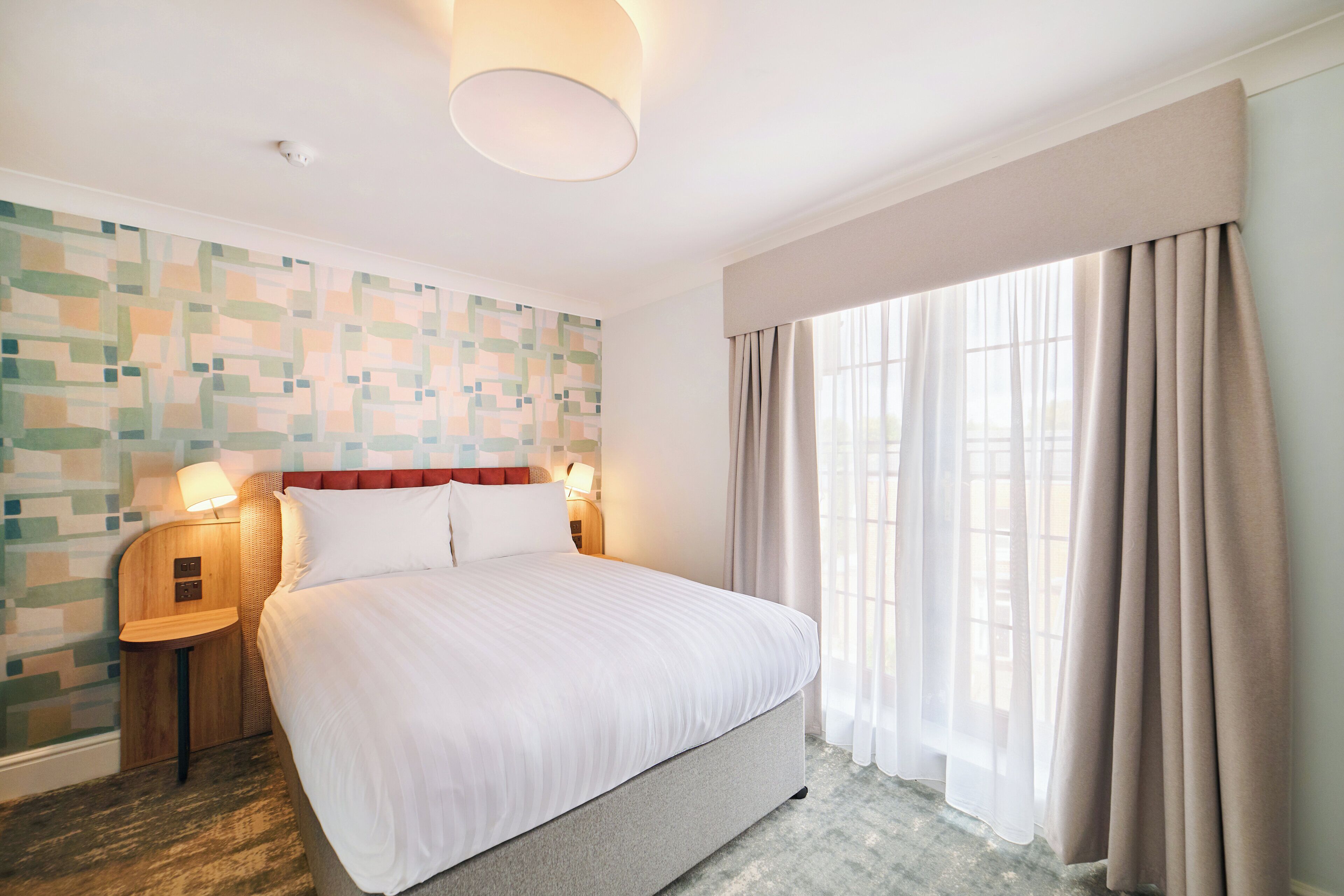 Une chambre d’hôtel avec un grand lit, deux lampes de chevet, un mur décoré d’un motif et une fenêtre avec des rideaux.