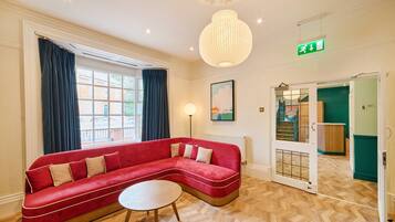 A red sofa with a round table in a room with wooden flooring, a large window with curtains, a floor lamp, and a green exit sign.