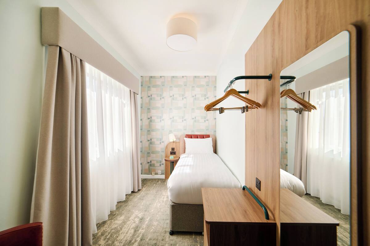 Una habitación de hotel con cama, mesita de noche, espejo y perchero.