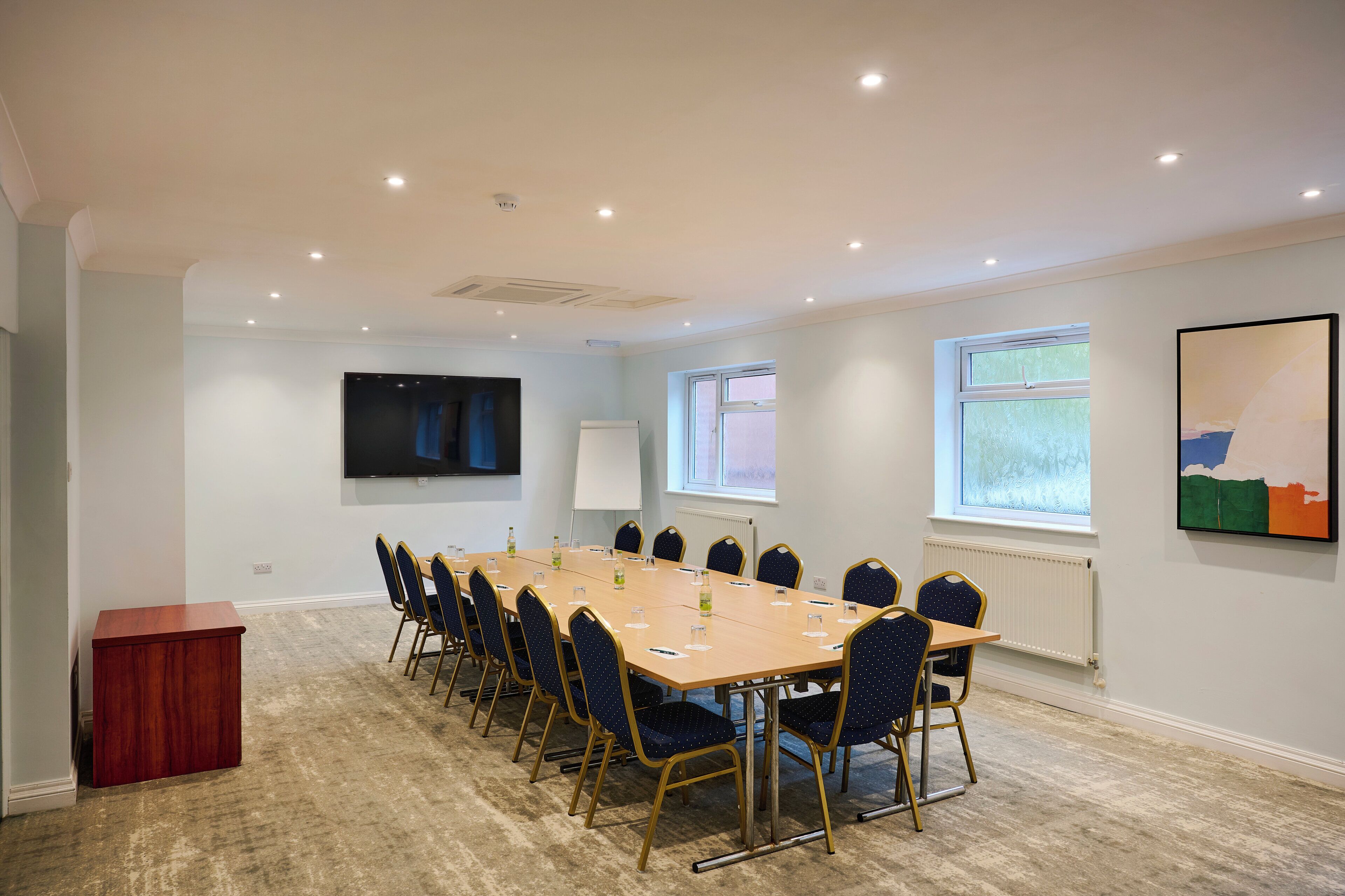A conference room with a long table, chairs, a podium, a TV, windows, and artwork.
