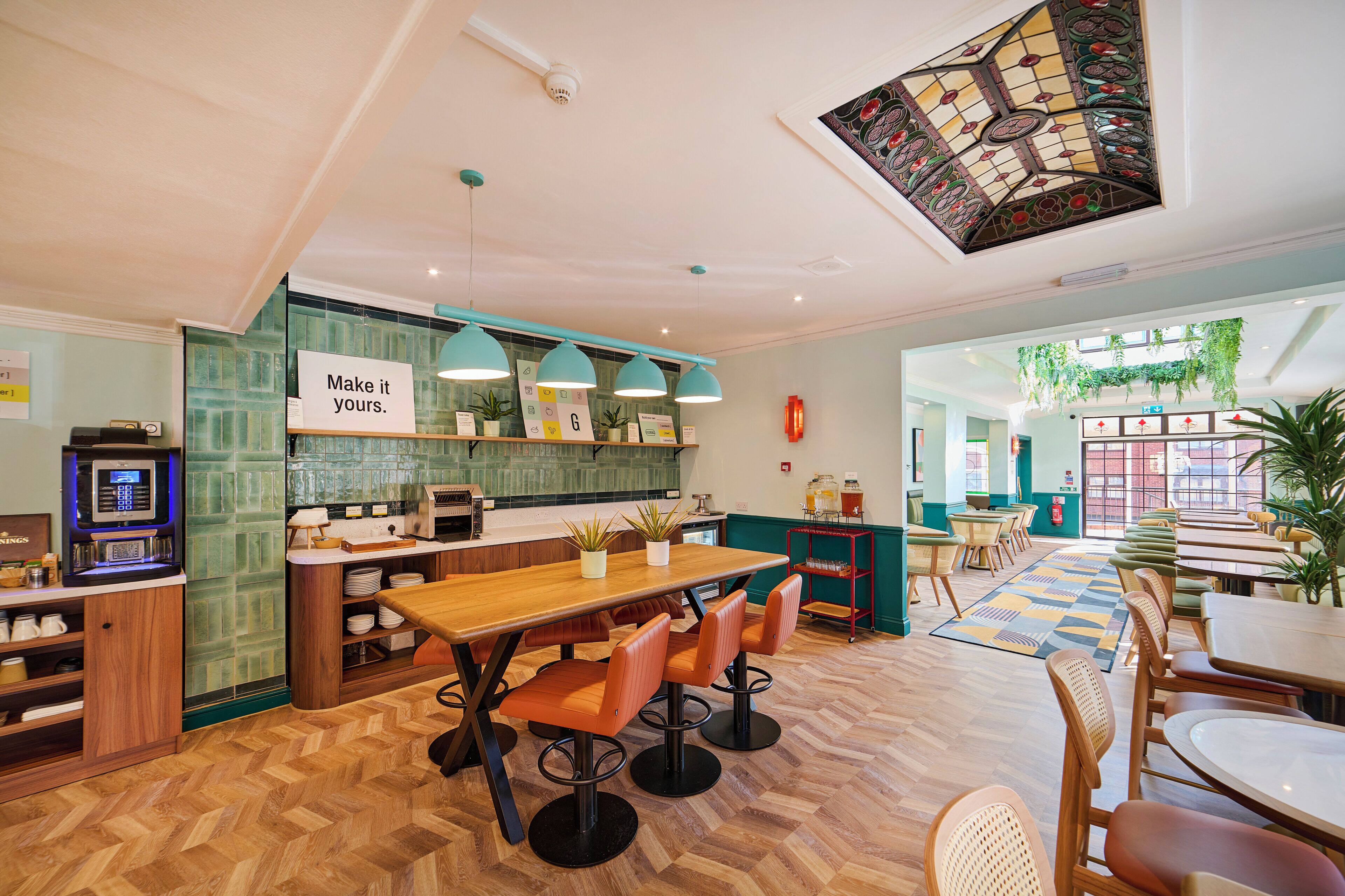 A modern restaurant with a wooden herringbone floor, a central dining table with orange chairs, and a green wall with a 'Make it yours' sign.