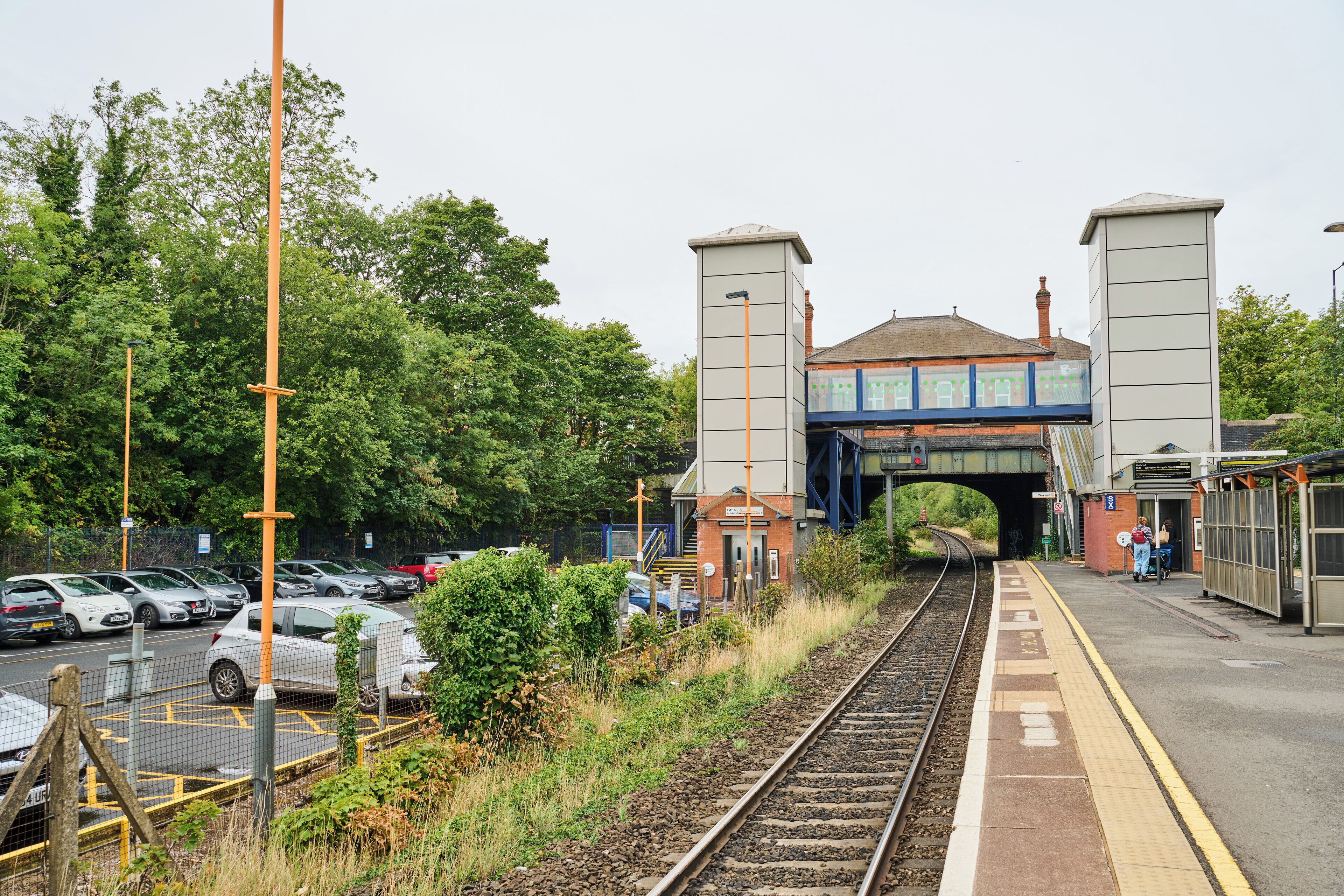 A railway station platform with a pedestrian bridge overhead, surrounded by parked cars and greenery.