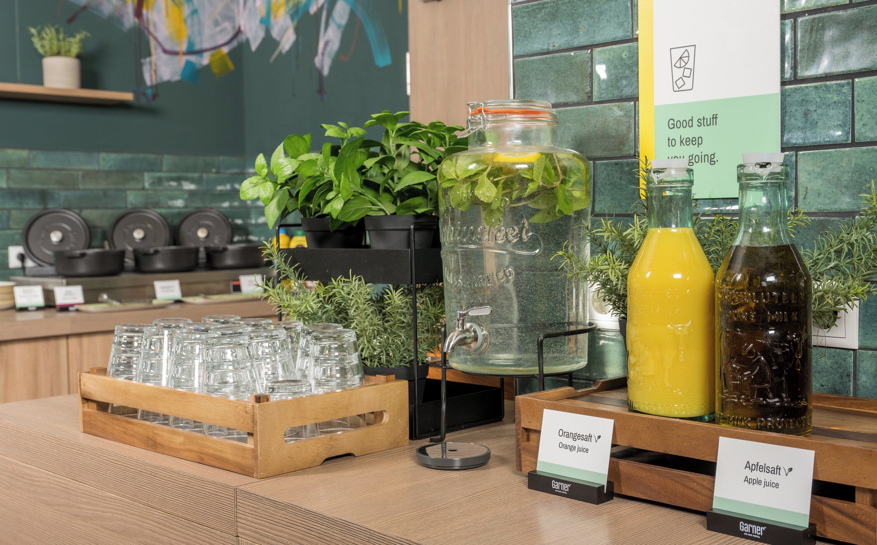 A beverage station with glass jars and bottles of orange and apple juice, surrounded by potted plants and a tiled backsplash.