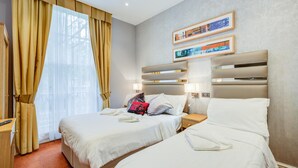 Ein Hotelzimmer mit zwei Betten, einem großen Fenster mit Vorhängen, einem Bettgestell mit Ablagen und gerahmten Bildern an der Wand.