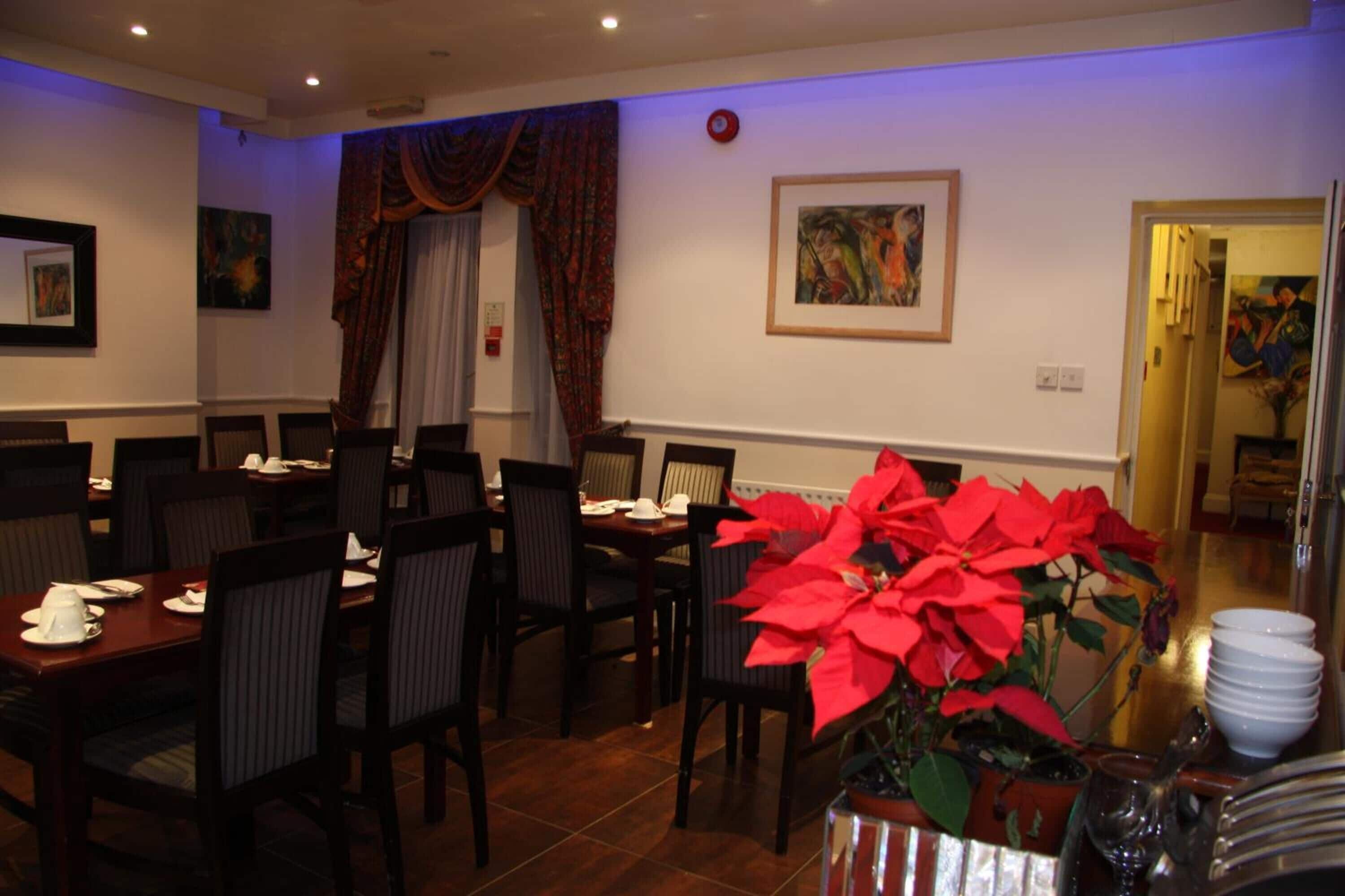 Un espace repas avec des tables dressées, une grande plante en pot ornée de fleurs rouges et des œuvres d’art accrochées aux murs.