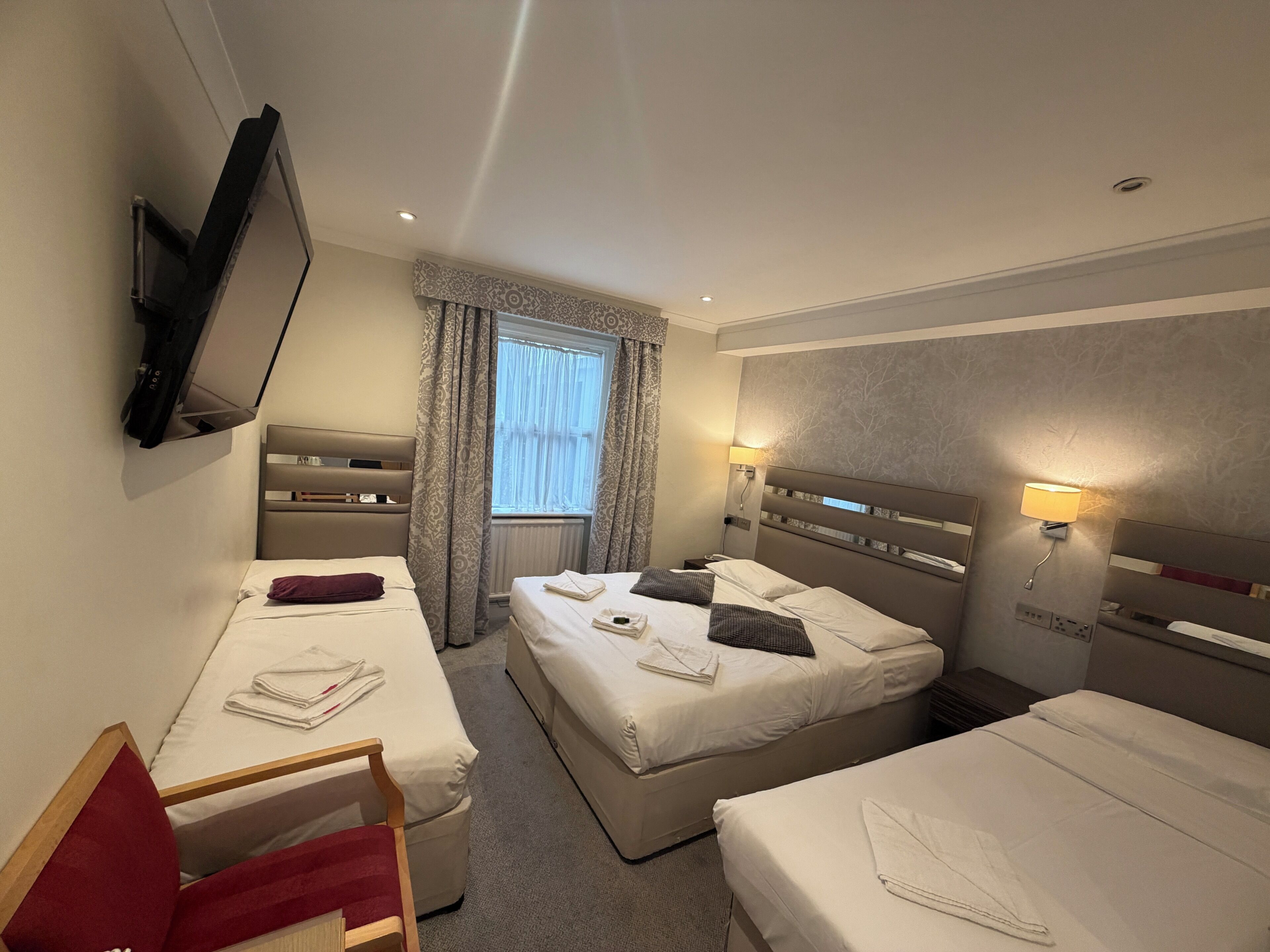 Ein Hotelzimmer mit zwei Einzelbetten, einem an der Wand montierten Fernseher, einem Fenster mit Vorhängen und Nachttischlampen.
