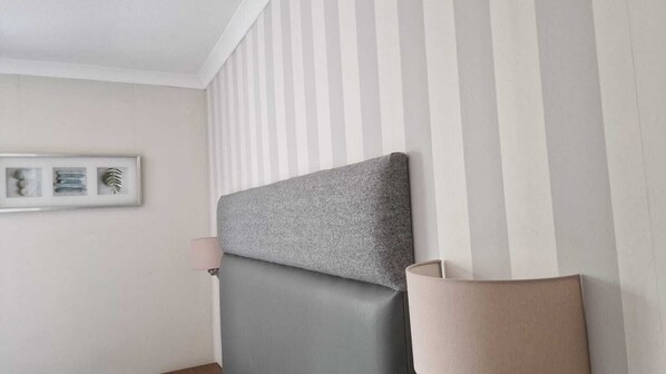 Una habitación con cabecera tapizada en gris, una lámpara de noche y un cuadro en la pared.