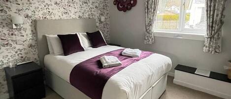 Une chambre à coucher bien aménagée, avec un grand lit, une table de chevet, une commode, une fenêtre avec des rideaux et un papier peint à motifs floraux.