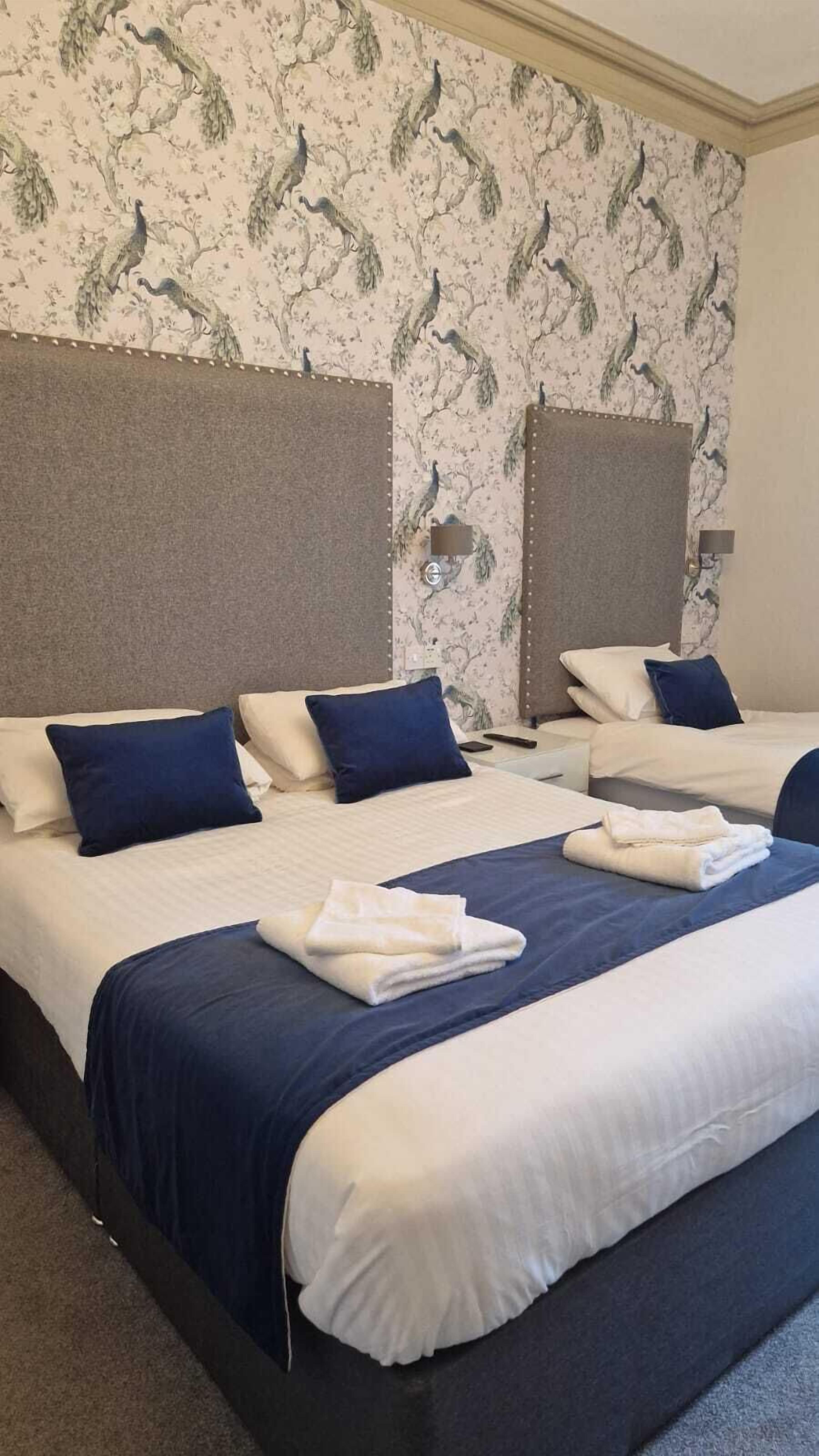 A hotel room with two beds, each with white linens and blue accents, and a patterned wallpaper featuring birds.